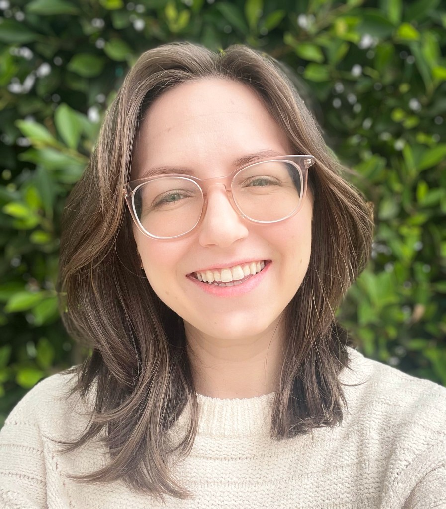 Rachel smiles at the camera. She is in a white knit sweater and wears translucent pink glasses. Her short hair is light brown and wavy. She is against a background of green leaves.